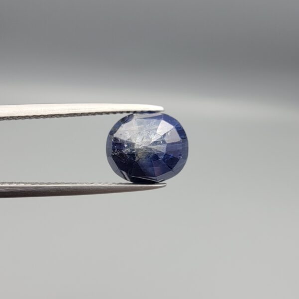 High-resolution macro photographs of a 5.40-carat natural Afghan Blue Indigo Sapphire (Neelam) held in steel gem tweezers, showcasing a masterfully faceted oval silhouette, deep midnight indigo-blue body color, and natural internal corundum silk structures sourced from Afghanistan.