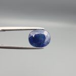 High-resolution macro photograph of a 3.76-carat natural Afghan Blue Indigo Sapphire (Neelam) held in steel gem tweezers, showcasing a finely faceted oval cut, deep indigo-blue body color, and natural corundum silk features sourced from Afghanistan.