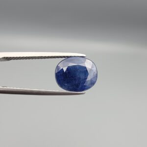 High-resolution macro photograph of a 3.76-carat natural Afghan Blue Indigo Sapphire (Neelam) held in steel gem tweezers, showcasing a finely faceted oval cut, deep indigo-blue body color, and natural corundum silk features sourced from Afghanistan.