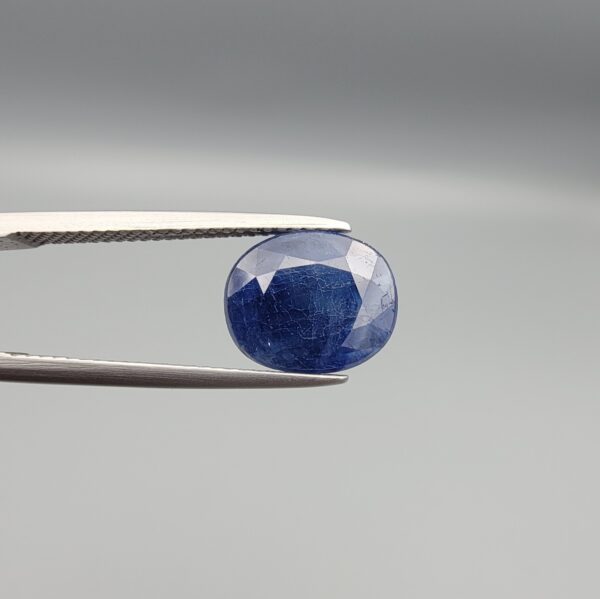 High-resolution macro photograph of a 3.76-carat natural Afghan Blue Indigo Sapphire (Neelam) held in steel gem tweezers, showcasing a finely faceted oval cut, deep indigo-blue body color, and natural corundum silk features sourced from Afghanistan.