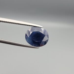 High-resolution macro photograph of a 4.34-carat natural Afghan Blue Indigo Sapphire (Neelam) held in steel gem tweezers, showcasing a finely faceted oval cut, deep indigo-blue body color, and natural internal corundum silk features sourced from Afghanistan.
