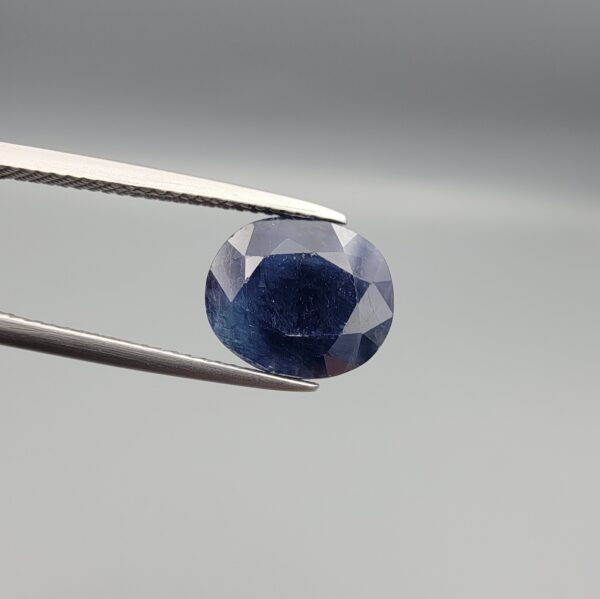 High-resolution macro photograph of a 4.34-carat natural Afghan Blue Indigo Sapphire (Neelam) held in steel gem tweezers, showcasing a finely faceted oval cut, deep indigo-blue body color, and natural internal corundum silk features sourced from Afghanistan.
