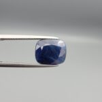 High-resolution macro photographs of a 7.31-carat natural Afghan Blue Indigo Sapphire (Neelam) held in steel gem tweezers, showcasing a soft cushion facet cut, deep indigo-blue body color, and natural internal corundum silk structures sourced from Afghanistan.