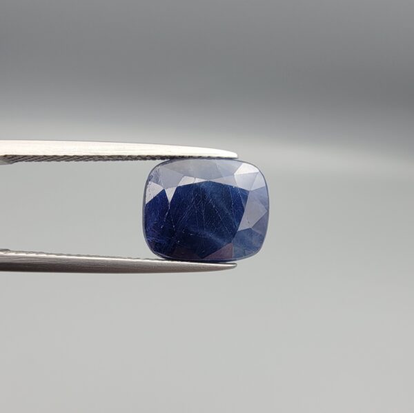High-resolution macro photographs of a 7.31-carat natural Afghan Blue Indigo Sapphire (Neelam) held in steel gem tweezers, showcasing a soft cushion facet cut, deep indigo-blue body color, and natural internal corundum silk structures sourced from Afghanistan.