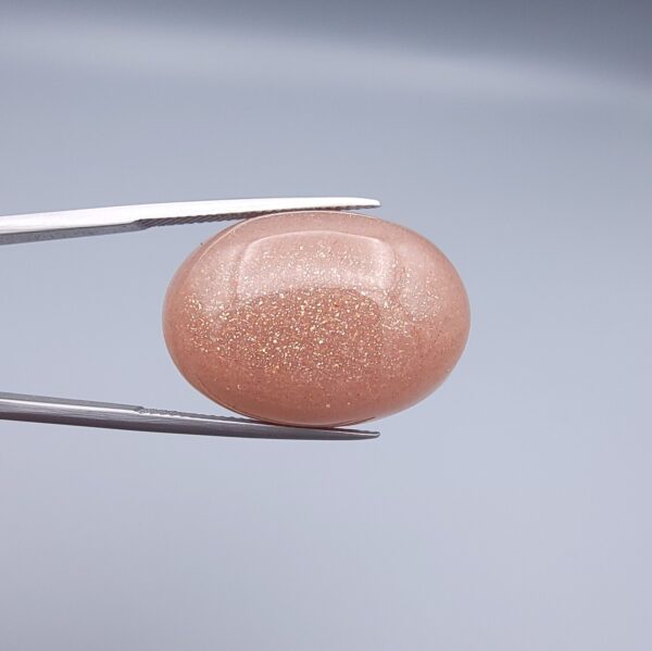 High-resolution macro photographs of a 34.71-carat natural Sunstone (Sang-e-Aftabi) held in steel tweezers, showcasing a smooth oval cabochon cut, warm peach-orange matrix, and dense aventurescent metallic specks that create a brilliant shimmering schiller effect.