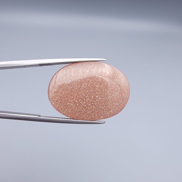 High-resolution macro photographs of a 34.71-carat natural Sunstone (Sang-e-Aftabi) held in steel tweezers, showcasing a smooth oval cabochon cut, warm peach-orange matrix, and dense aventurescent metallic specks that create a brilliant shimmering schiller effect.