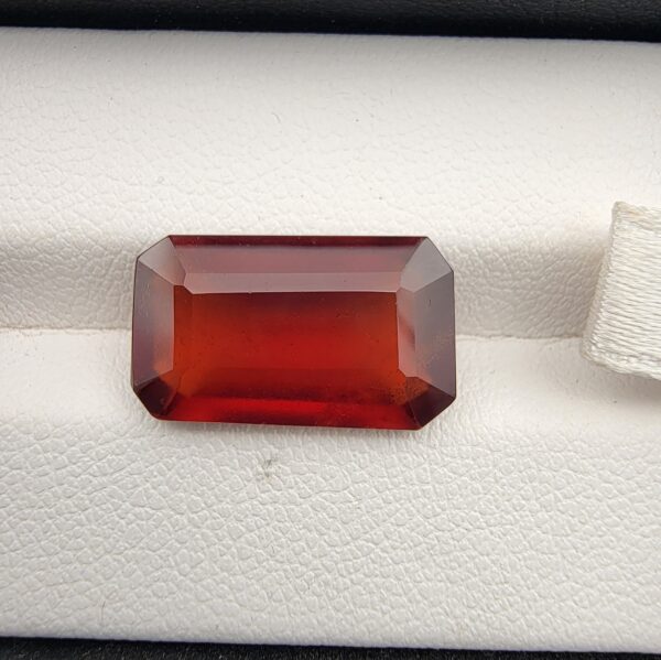 High-resolution macro photographs of a 9.59-carat natural Hessonite Garnet (Gomed) resting on a white leather display cushion inside a black leatherette gem box, displaying an emerald step-cut rectangular silhouette, rich reddish-brown to warm honey-amber color, and characteristic internal heat-wave swirl effect (treacle).