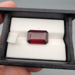 High-resolution macro photographs of a 10.00-carat natural Hessonite Garnet (Gomed) displayed on a white leather cushion inside a black presentation box, showcasing a clean emerald step-cut rectangular silhouette, rich reddish-brown to warm honey-amber body color, and characteristic internal heat-wave swirl structure (treacle effect).