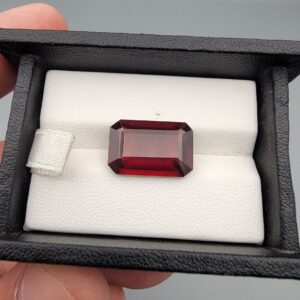 High-resolution macro photographs of a 10.00-carat natural Hessonite Garnet (Gomed) displayed on a white leather cushion inside a black presentation box, showcasing a clean emerald step-cut rectangular silhouette, rich reddish-brown to warm honey-amber body color, and characteristic internal heat-wave swirl structure (treacle effect).