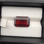 High-resolution macro photographs of a 12.00-carat natural Hessonite Garnet (Gomed) displayed inside a black leatherette display box and placed on the back of a hand for scale reference, showcasing a clean step-cut rectangular emerald silhouette, rich reddish-brown to warm cinnamon-amber hues, and characteristic internal heat-wave swirl transparency (treacle effect).
