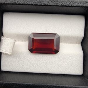 High-resolution macro photographs of a 12.00-carat natural Hessonite Garnet (Gomed) displayed inside a black leatherette display box and placed on the back of a hand for scale reference, showcasing a clean step-cut rectangular emerald silhouette, rich reddish-brown to warm cinnamon-amber hues, and characteristic internal heat-wave swirl transparency (treacle effect).