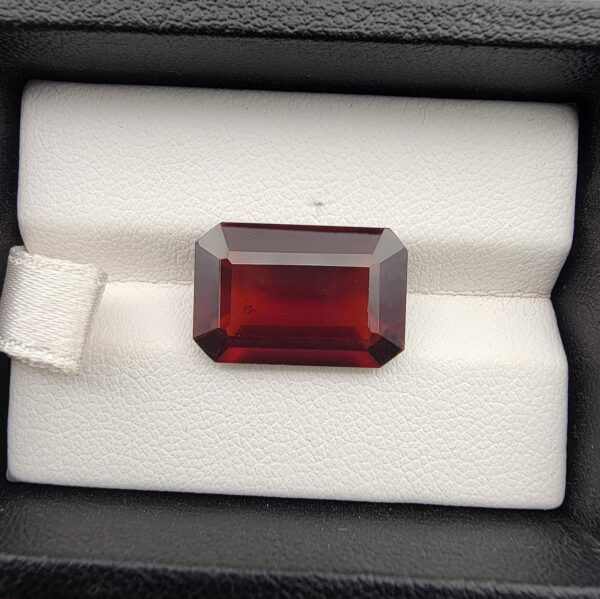 High-resolution macro photographs of a 12.00-carat natural Hessonite Garnet (Gomed) displayed inside a black leatherette display box and placed on the back of a hand for scale reference, showcasing a clean step-cut rectangular emerald silhouette, rich reddish-brown to warm cinnamon-amber hues, and characteristic internal heat-wave swirl transparency (treacle effect).