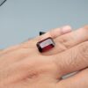High-resolution macro photographs of a 12.00-carat natural Hessonite Garnet (Gomed) displayed inside a black leatherette display box and placed on the back of a hand for scale reference, showcasing a clean step-cut rectangular emerald silhouette, rich reddish-brown to warm cinnamon-amber hues, and characteristic internal heat-wave swirl transparency (treacle effect).