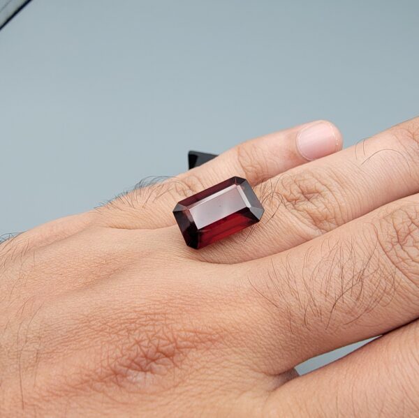 High-resolution macro photographs of a 12.00-carat natural Hessonite Garnet (Gomed) displayed inside a black leatherette display box and placed on the back of a hand for scale reference, showcasing a clean step-cut rectangular emerald silhouette, rich reddish-brown to warm cinnamon-amber hues, and characteristic internal heat-wave swirl transparency (treacle effect).