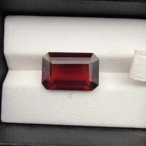 High-resolution macro photographs of an 11.32-carat natural Hessonite Garnet (Gomed) displayed on a white textured leather cushion inside a black display box, showcasing a precision emerald step-cut rectangular silhouette, rich deep reddish-brown to warm cinnamon-amber hues, and signature internal heat-wave transparency (treacle effect).
