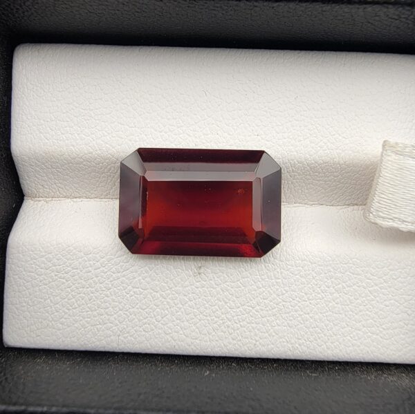 High-resolution macro photographs of an 11.32-carat natural Hessonite Garnet (Gomed) displayed on a white textured leather cushion inside a black display box, showcasing a precision emerald step-cut rectangular silhouette, rich deep reddish-brown to warm cinnamon-amber hues, and signature internal heat-wave transparency (treacle effect).