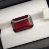 High-resolution macro photographs of an 11.32-carat natural Hessonite Garnet (Gomed) displayed on a white textured leather cushion inside a black display box, showcasing a precision emerald step-cut rectangular silhouette, rich deep reddish-brown to warm cinnamon-amber hues, and signature internal heat-wave transparency (treacle effect).