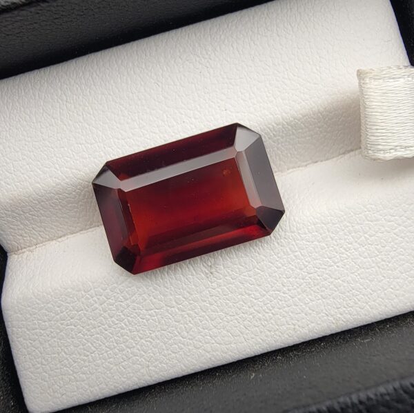 High-resolution macro photographs of an 11.32-carat natural Hessonite Garnet (Gomed) displayed on a white textured leather cushion inside a black display box, showcasing a precision emerald step-cut rectangular silhouette, rich deep reddish-brown to warm cinnamon-amber hues, and signature internal heat-wave transparency (treacle effect).