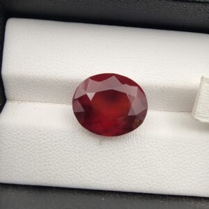High-resolution macro photograph of a 9.92-carat natural Hessonite Garnet (Gomed) resting on a white textured leather display bed inside a black display box, showcasing a masterfully faceted oval silhouette, deep reddish-brown to warm honey-amber color, and characteristic internal heat-wave swirl transparency (treacle effect).