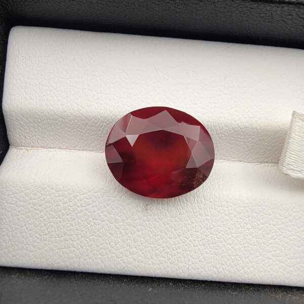 High-resolution macro photograph of a 9.92-carat natural Hessonite Garnet (Gomed) resting on a white textured leather display bed inside a black display box, showcasing a masterfully faceted oval silhouette, deep reddish-brown to warm honey-amber color, and characteristic internal heat-wave swirl transparency (treacle effect).