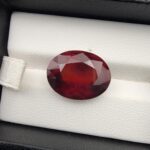 High-resolution macro photograph of a 13.76-carat natural Hessonite Garnet (Gomed) resting on a white leather display bed inside a black display box, showcasing a masterfully faceted oval silhouette, deep reddish-brown to warm honey-amber color, and characteristic internal heat-wave swirl transparency (treacle effect).