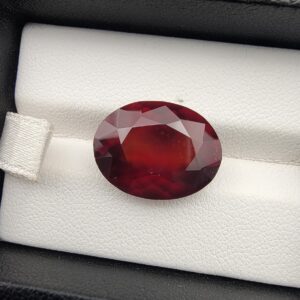 High-resolution macro photograph of a 13.76-carat natural Hessonite Garnet (Gomed) resting on a white leather display bed inside a black display box, showcasing a masterfully faceted oval silhouette, deep reddish-brown to warm honey-amber color, and characteristic internal heat-wave swirl transparency (treacle effect).