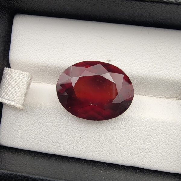 High-resolution macro photograph of a 13.76-carat natural Hessonite Garnet (Gomed) resting on a white leather display bed inside a black display box, showcasing a masterfully faceted oval silhouette, deep reddish-brown to warm honey-amber color, and characteristic internal heat-wave swirl transparency (treacle effect).