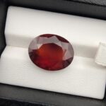 High-resolution macro photograph of a 13.76-carat natural Hessonite Garnet (Gomed) displayed on a white textured leather bed inside a black box, showcasing a finely faceted oval cut, deep reddish-brown to warm honey-amber body color, and characteristic internal heat-wave swirl transparency (treacle effect).