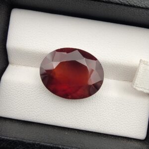 High-resolution macro photograph of a 13.76-carat natural Hessonite Garnet (Gomed) displayed on a white textured leather bed inside a black box, showcasing a finely faceted oval cut, deep reddish-brown to warm honey-amber body color, and characteristic internal heat-wave swirl transparency (treacle effect).