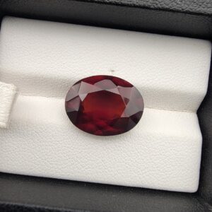 High-resolution macro photograph of a 10.38-carat natural Hessonite Garnet (Gomed) resting on a white leather display cushion inside a black display box, showcasing a finely faceted oval cut, deep reddish-brown to warm honey-amber body color, and characteristic internal heat-wave swirl transparency (treacle effect).