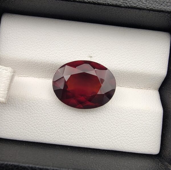 High-resolution macro photograph of a 10.38-carat natural Hessonite Garnet (Gomed) resting on a white leather display cushion inside a black display box, showcasing a finely faceted oval cut, deep reddish-brown to warm honey-amber body color, and characteristic internal heat-wave swirl transparency (treacle effect).