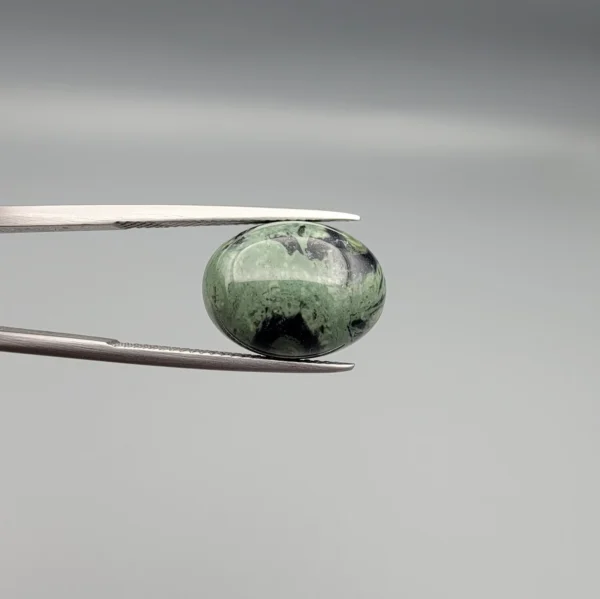High-resolution macro photographs showing a 10.00-carat natural Serpentine (Zehr Mora) oval cabochon held in gemological tweezers from front and back views, displaying a sage-green body matrix with striking black mineral inclusions and veining under a smooth dome polish.