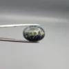 High-resolution macro photographs showing a 12.97-carat natural Serpentine (Zehr Mora) oval cabochon held in gemological tweezers from front and back views, displaying a dark charcoal-green matrix with distinct pale-green circular orbital "eye" inclusions under a high-luster polished dome.