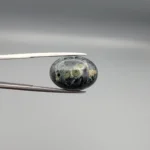 High-resolution macro photographs showing a 12.97-carat natural Serpentine (Zehr Mora) oval cabochon held in gemological tweezers from front and back views, displaying a dark charcoal-green matrix with distinct pale-green circular orbital "eye" inclusions under a high-luster polished dome.