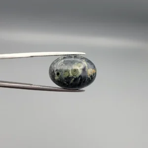 High-resolution macro photographs showing a 12.97-carat natural Serpentine (Zehr Mora) oval cabochon held in gemological tweezers from front and back views, displaying a dark charcoal-green matrix with distinct pale-green circular orbital "eye" inclusions under a high-luster polished dome.