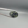 High-resolution macro photographs showing a 12.97-carat natural Serpentine (Zehr Mora) oval cabochon held in gemological tweezers from front and back views, displaying a dark charcoal-green matrix with distinct pale-green circular orbital "eye" inclusions under a high-luster polished dome.