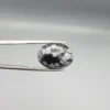 High-resolution macro photographs showing a 13.78-carat natural Serpentine (Zehr Mora) oval cabochon held in tweezers and showcased on hand, displaying a deep jet-black base matrix contrasting with crisp white snowflake-like inclusions under a high-luster polished dome.