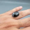 High-resolution macro photographs showing a 13.78-carat natural Serpentine (Zehr Mora) oval cabochon held in tweezers and showcased on hand, displaying a deep jet-black base matrix contrasting with crisp white snowflake-like inclusions under a high-luster polished dome.