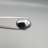 High-resolution macro photograph showing a 19.71-carat natural Hematite (Sang-E-Hadeed) oval cabochon held in gemological tweezers, exhibiting a mirror-like metallic luster, deep gunmetal grey sheen, and a perfectly polished dome.