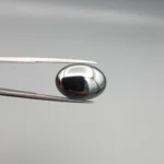 High-resolution macro photograph showing a 19.71-carat natural Hematite (Sang-E-Hadeed) oval cabochon held in gemological tweezers, exhibiting a mirror-like metallic luster, deep gunmetal grey sheen, and a perfectly polished dome.