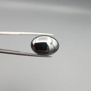 High-resolution macro photograph showing a 19.71-carat natural Hematite (Sang-E-Hadeed) oval cabochon held in gemological tweezers, exhibiting a mirror-like metallic luster, deep gunmetal grey sheen, and a perfectly polished dome.