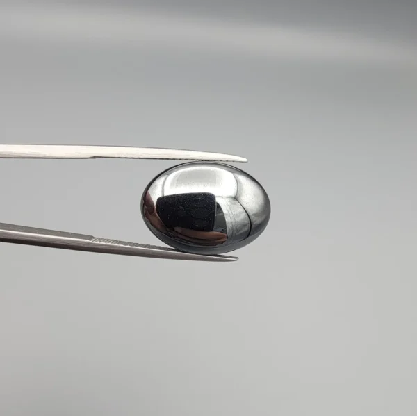 High-resolution macro photograph showing a 19.71-carat natural Hematite (Sang-E-Hadeed) oval cabochon held in gemological tweezers, exhibiting a mirror-like metallic luster, deep gunmetal grey sheen, and a perfectly polished dome.