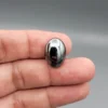 High-resolution macro photograph showing a 19.71-carat natural Hematite (Sang-E-Hadeed) oval cabochon held in gemological tweezers, exhibiting a mirror-like metallic luster, deep gunmetal grey sheen, and a perfectly polished dome.