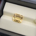 High-resolution macro photograph displaying a 4.42-carat natural Citrine Quartz (Sunela) octagon step cut gemstone resting in a white display box, showcasing crisp emerald-cut step facets, high transparency, and a refined golden-yellow color.