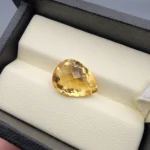 High-resolution macro photograph showing a 5.26-carat natural Citrine Quartz (Sunela) pear cut gemstone with checkerboard crown faceting resting in a luxury white-padded display box, exhibiting brilliant golden-yellow color and crystal clarity.