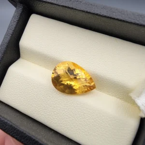 High-resolution macro photograph displaying a 4.88-carat natural Citrine Quartz (Sunela) pear cut faceted gemstone set in a luxury white-padded display box, showing intense golden-honey saturation, brilliant light refraction, and high crystal transparency.