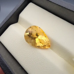 High-resolution macro photographs showing a 6.57-carat natural Citrine Quartz (Sunela) pear cut faceted gemstone in a white-padded display box and held in gemological tweezers, exhibiting brilliant golden-yellow color, high crystal transparency, and crisp precision faceting.