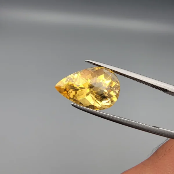 High-resolution macro photographs showing a 6.57-carat natural Citrine Quartz (Sunela) pear cut faceted gemstone in a white-padded display box and held in gemological tweezers, exhibiting brilliant golden-yellow color, high crystal transparency, and crisp precision faceting.