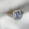 Close-up photograph of an 18K gold ring featuring a 1.53-carat emerald-cut pastel blue natural Ceylon sapphire centerpiece surrounded by a shimmering halo of 0.33-carat natural diamond accents against a soft white fabric background.