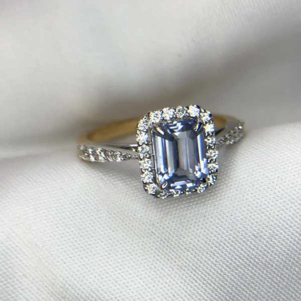 Close-up photograph of an 18K gold ring featuring a 1.53-carat emerald-cut pastel blue natural Ceylon sapphire centerpiece surrounded by a shimmering halo of 0.33-carat natural diamond accents against a soft white fabric background.
